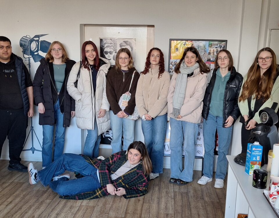 Das Bild zeigt eine Gruppe von elf Schülern in einem Raum mit Holzboden und wei-ßen Wänden. Zehn Personen stehen und eine liegt. Die meisten Personen tragen winterliche Kleidung. Die Gruppe ist gemischt, mit mehreren Frauen und zwei Män-nern auf der linken Seite. Die Gruppe steht entspannt zusammen, einige lächeln leicht. Die Person in der Mitte des Vordergrunds liegt auf dem Boden und stützt sich auf einen Ellenbogen. Die Stimmung wirkt locker und freundlich.